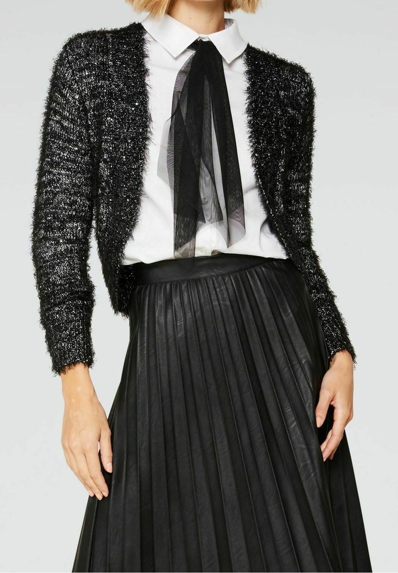 Cardigan noir en tricot avec une texture duveteuse, associé à une chemise blanche ornée d'un nœud papillon transparent noir et une jupe plissée noire.