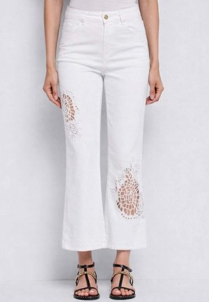 Persona che indossa pantaloni bianchi a campana corti con motivi traforati in pizzo sulle cosce e sandali neri con dettagli in catena dorata.