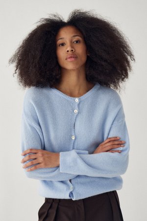 Ženska z bujnimi kodrastimi lasmi nosi svetlo modro srajčno jopico in črne hlače, stoji z rokami prekrižanimi.