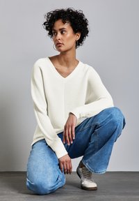 Maglione bianco a V in tessuto abbinato a jeans boyfriend blu e sneakers beige, con un focus su un look informale e morbido.