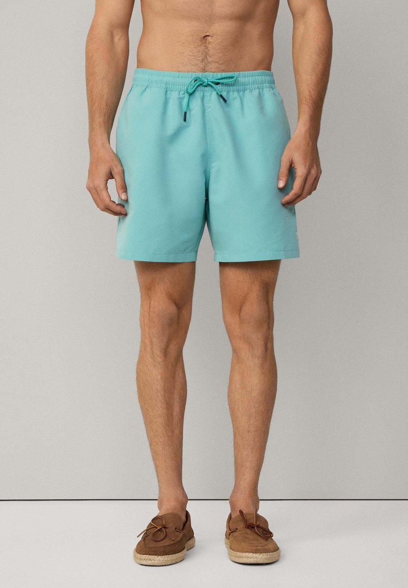 Boxer da bagno azzurro chiaro con vita elastica e cordoncino, texture liscia. Scarpe slip-on in suede marrone con dettagli in corda.