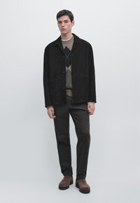 Jeune homme portant une veste noire à boutons, un pull à losanges, un pantalon sombre et des bottes en daim marron, debout devant un fond clair uni.