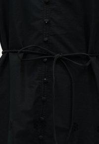 Black garment with fabric-covered buttons down center and a tied belt at the waist, made of lightweight, slightly textured fabric.