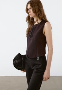 Mouwloze donkerpaarse top met verticale zwarte details en knopen. Gecombineerd met zwarte broek en een geweven zwarte handtas.