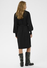 Crna pletena haljina s dugim napuhanim rukavima i vezanom strukom. Model nosi crne visokokvalitetne čizme do koljena s kopčama. Jednostavan, elegantan dizajn.