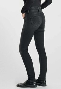 Jean en denim noir avec une coupe ajustée, une taille haute et des coutures apparentes. Deux poches arrière avec un détail de couture incurvé et une texture lisse.