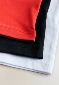 Kolm puuvillast T-särki virnastatult: punane ülal, must keskel ja valge all. Kõigil on soonikuga kaelus ning sirged õmblused.