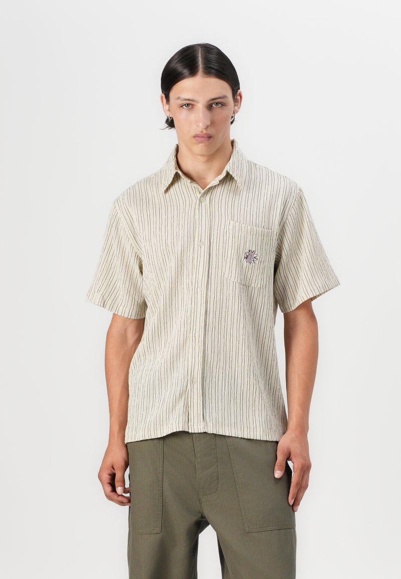 Kortermet, beige skjorte med vertikale striper, knapping foran og en brystlomme med et lite brodert design.