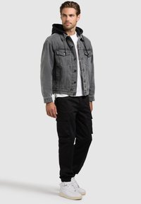 Homme debout portant une veste en jean grise avec capuche noire, t-shirt blanc, pantalon cargo noir et baskets blanches, regardant vers la droite.