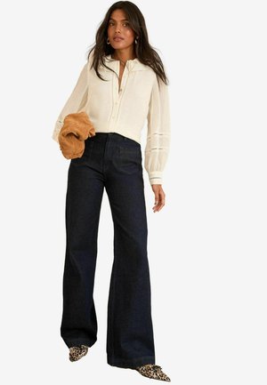 Blusa bianca a maniche lunghe con bottoni frontali, jeans a vita alta blu scuro a campana e scarpe con stampa leopardata. Modella che tiene un accessorio marrone.