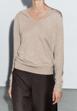 Person wearing beige V-neck long-sleeve sweater and dark brown pants, standing against a plain light background.