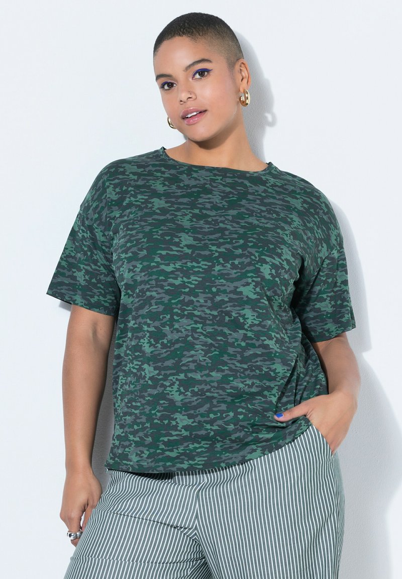 Person mit kurzen Haaren, der ein grünes Tarn-T-Shirt und weiße Hosen mit Streifen trägt und mit einer Hand in der Tasche vor weißem Hintergrund posiert.