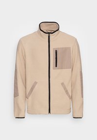 Giacca in pile crema con colletto alto, chiusura con zip nera, tasca sul petto crema con zip, e toppe crema sui gomiti e tasche laterali.