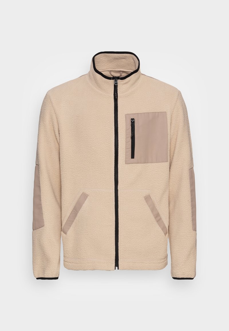 Krämfärgad fleecjacka med ståkrage, svart dragkedja, beige bröstficka med dragkedja, samt beige armbågsfläckar och sidfickor.