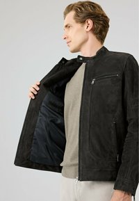 Zwarte suède jas met een rechtopstaande kraag, ritszakken en een gladde satijnen voering, gepresenteerd met een gedeeltelijk open voorkant.