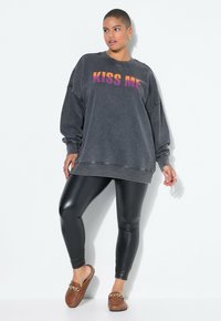 Sudadera negra de gran tamaño con "KISS ME" en un degradado de naranja a púrpura. Combinada con leggings negros brillantes y zuecos marrones con un detalle de cadena.
