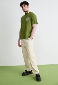 T-shirt en coton vert olive à manches courtes avec une poche avant, associé à un pantalon beige clair et des chaussures noires à semelles épaisses.