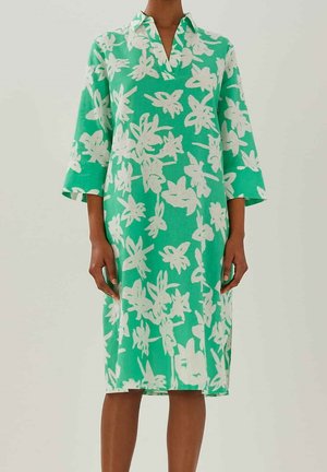 Kvinde iført en grøn knælængde kjole med hvide blomsterprint og trekvartærmede, stående mod en ensfarvet baggrund.