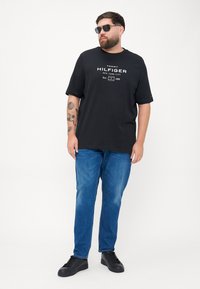 Czarny t-shirt z krótkim rękawem z białym napisem, zestawiony z niebieskimi dżinsami i czarnymi sneakersami. Dodatki to ciemne okulary przeciwsłoneczne.