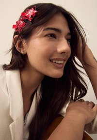 Red sequin hair clips shaped like stars adorn dark hair, with a cream-colored blazer and soft brown fabric in the background.