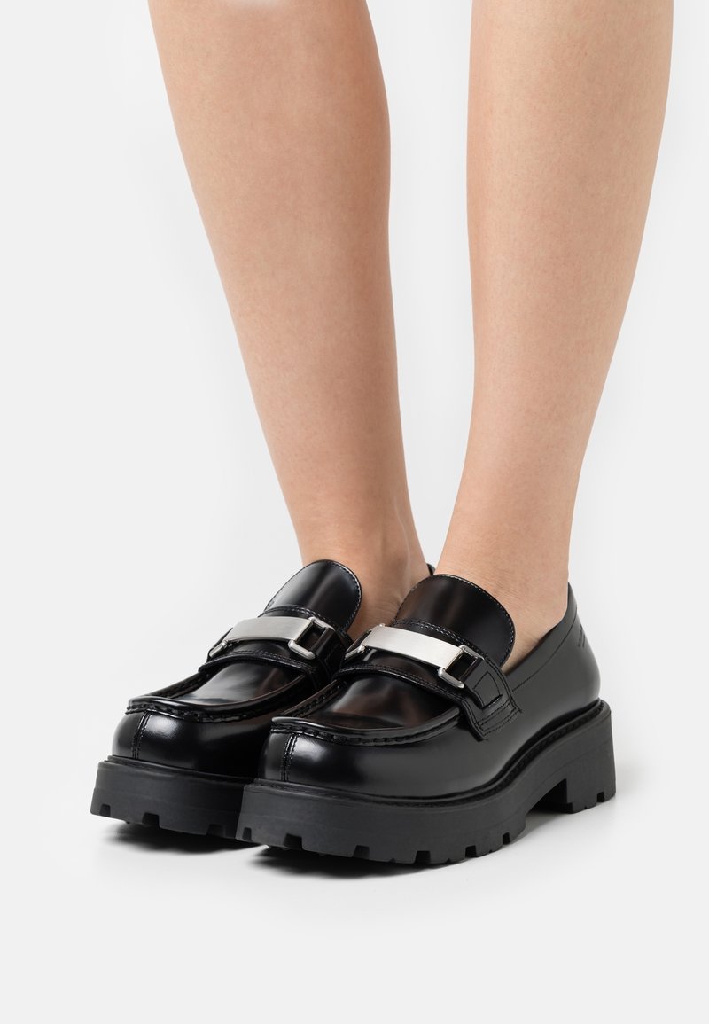 Mocassins en cuir noir avec une finition brillante, dotés d'une boucle argentée audacieuse et d'une semelle noire épaisse à détail de crampons.