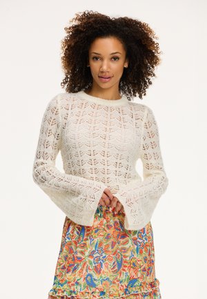 Maglione bianco lavorato a maglia con maniche a campana e motivo in pizzo. La modella indossa una gonna floreale multicolore con orlo arricciato.