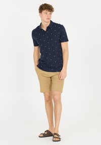 Marinblå polo skjorta med vita mönster, korta ärmar och krage. Kombineras med ljusa khaki-shorts och bruna sandaler. Slät bomullsmaterial.