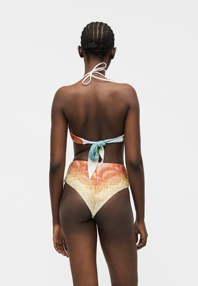 Femme aux cheveux tressés vue de dos, portant un haut de bikini multicolore à col montant et un bas de bikini taille haute sur un fond uni.