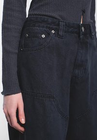 Tummat farkut, joissa on korkea vyötärö, suuret sivutaskut, hopeiset yksityiskohdat ja sileä pinta. Läheltä otettu kuva kädestä, joka lepää kankaan päällä.