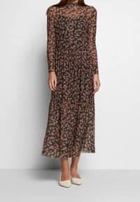 Langes schwarzes Kleid aus transparenter Stoff, mit floralem Muster in Rosatönen, Rundhals und gerafftem Bund; kombiniert mit weißen Schuhen.