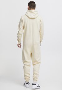 Overall in morbido tessuto color crema con cappuccio, vestibilità comoda, maniche lunghe, polsini a coste e una tasca sul retro per riporre oggetti.