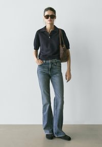 Marineblaues Poloshirt, hellblaue Schlagjeans, schwarze flache Schuhe und braune Handtasche. Das Model trägt übergroße Sonnenbrillen und steht vor einem schlichten Hintergrund.