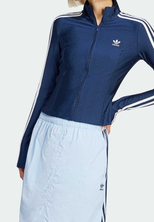 Vrouw draagt een marineblauw Adidas jas met rits en witte strepen en een lichtblauwe Adidas rok met een elastische tailleband.