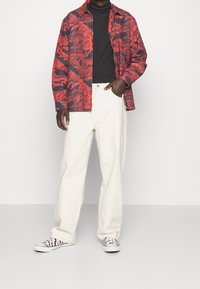 Giacca a righe rossa e nera sopra un dolcevita nero, abbinata a pantaloni larghi bianchi e sneakers con motivo bianco e nero.