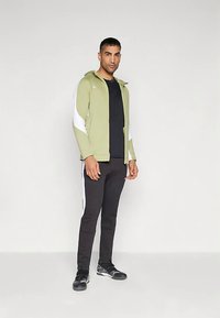 Giacca con cerniera verde oliva chiaro con accenti bianchi, abbinata a pantaloni neri aderenti e scarpe sportive nere, in piedi su uno sfondo neutro.