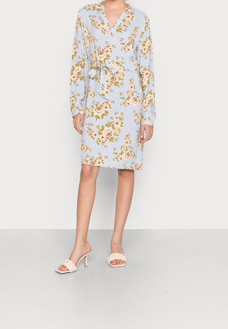 Robe florale bleu clair à manches longues, col en V et ceinture à nouer. Sandales blanches tressées à talons complètent la tenue.