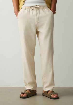 Beežid paelaga püksid, kantud oliivroheliste sandaalidega, inimene seisab käed taskutes ühevärvilise tausta ees.