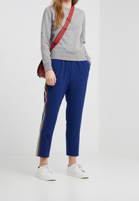 Persona vestida con pantalones azules con franjas laterales rojas, verdes y blancas, sudadera gris con logotipos de colores, zapatillas blancas y un bolso cruzado rojo.