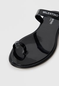 Black patent leather flip-flop sandal features a flat sole, T-shaped strap, and embossed logo on the band. Textured edge detailing.