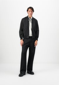 Black bomber jacket with two large front pockets, worn over a white shirt. Paired with black wide-leg trousers and chunky black shoes.
