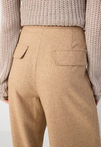 Tan broeken van een gestructureerde stof. Kenmerken zijn onder andere twee achterzakken met kleppen en een recht gesneden ontwerp. Gestyled met een gebreid top.
