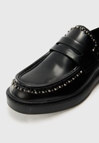 Mocassins en cuir noir au design épuré, ornés de clous en métal argenté le long du bord supérieur et d'une finition lisse et brillante. Semelle en caoutchouc.