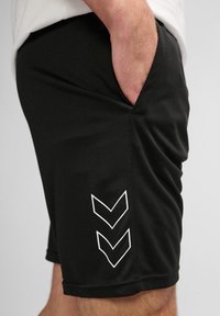 Osoba nosząca czarne spodenki z dwoma białymi logo w kształcie jodełki po boku, ręka częściowo w prawych kieszeniach.