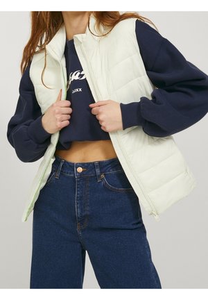 Frau in weißer gesteppter Weste über einem marineblauen Sweatshirt und hoch taillierter blauer Jeans, hält die Weste geöffnet, um den Bauch und Kleidungsdetails zu zeigen.