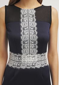 Marineblauwe jurk met een zwarte mesh halslijn, voorzien van witte kanten details in het midden en een kanten tailleband, gladde stof, nauwsluitende pasvorm.