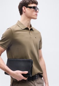 Νέος άνδρας που φοράει καφέ πόλο μπλούζα και μαύρα γυαλιά ηλίου Calvin Klein, κρατώντας μαύρο δερμάτινο clutch στην περιοχή της μέσης.