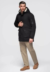 Parka negra con capucha, forrada interiormente con un suave tejido crema, con bolsillos delanteros y un bolsillo lateral con cremallera. Combinada con pantalones beige y botas marrones.