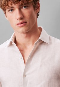 Νέος άνδρας με σγουρά μαλλιά που φοράει ανοιχτόχρωμο πουκάμισο με κουμπιά και ανοιχτό γιακά σε μονόχρωμο γκρι φόντο.