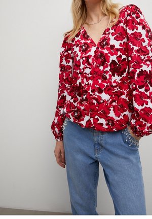 Femme portant un chemisier à manches longues rouge et blanc à motif floral avec des manches bouffantes et un jean bleu avec des détails en perles sur les poches.