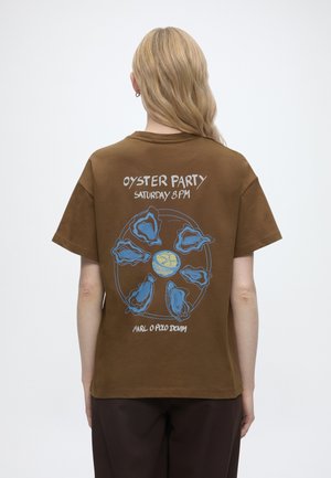 Person med langt blondt hår iført en brun T-shirt med et blåt østerskalsdesign og teksten "Oyster Party Saturday 8 PM" på bagsiden.